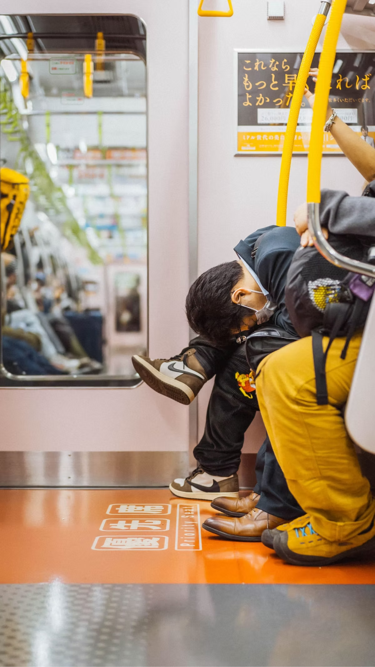 People sitting on the floor of a train, with some leaning forward and resting their heads on their knees. They are wearing masks and casual clothing, with one person in yellow pants and another in black sneakers. There is a mirror on the side and saf