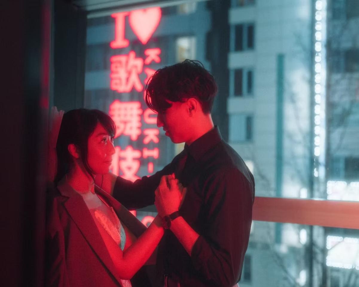 A couple stands closely in front of a neon "I Love Kabukicho" sign in the vibrant district of Tokyo at night, bathed in red lighting.