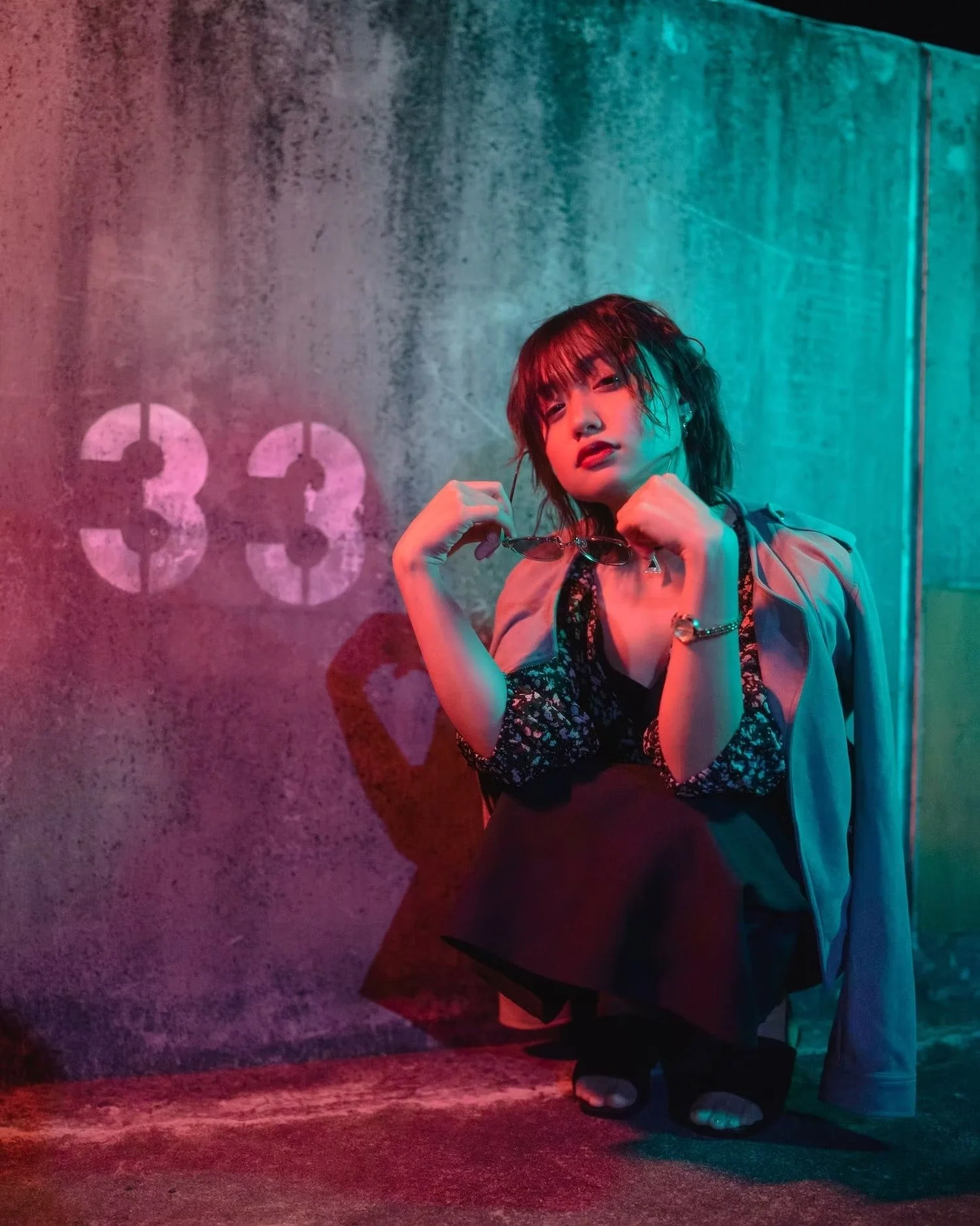 A woman with short dark hair and bangs, wearing a beige jacket and black clothing, is squatting while holding a pair of sunglasses. She is in a dimly lit room with multicolored neon lighting. The background features a concrete wall with the number '3