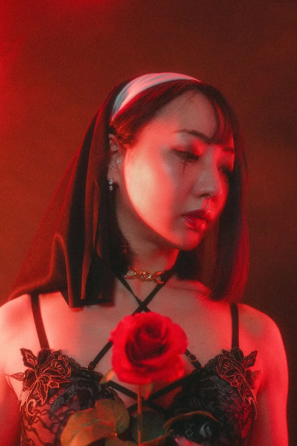 A woman with black hair wearing a black lace top, a choker, and jewelry, holding a red rose in front of her chest, with red lighting and a dark background.