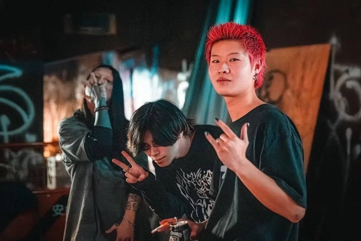 Three young people in a dimly lit room, with graffiti on the walls. One with bright red hair making a hand gesture, another with black hair making a peace sign, and the third person with dark hair holding a phone in Osaka, Namba area.