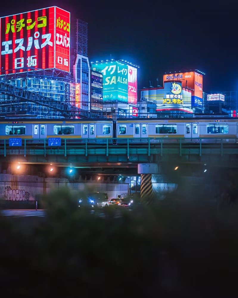 Night view of a bustling cityscape in Shinjuku with brightly lit buildings adorned with Japanese advertisements, a train crossing an elevated track, and cars on the road below.