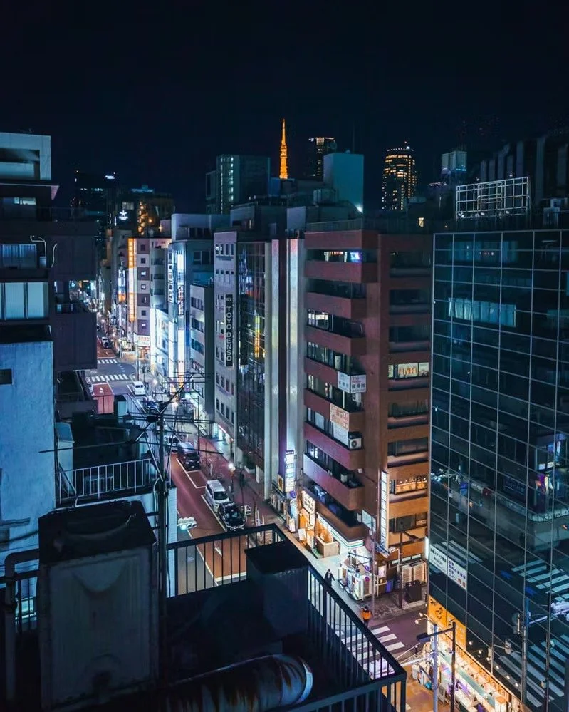 Nighttime cityscape of a busy urban street with illuminated buildings, cars, and signs, viewed from a higher floor from a rooftop in Shimbashi, Tokyo.