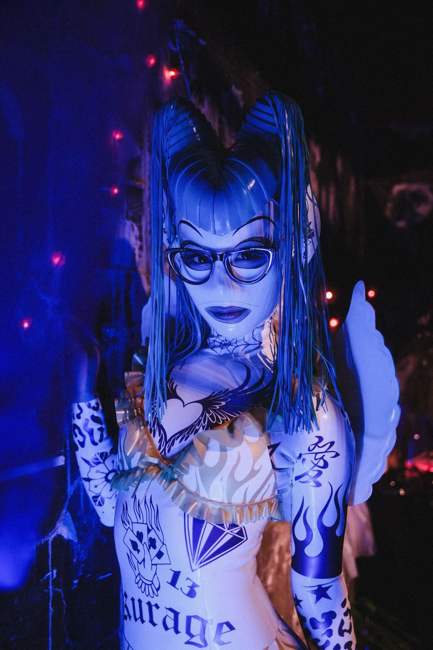 Bondage Ball 
---
A person in a large, colorful costume with a mask and glasses, illuminated by blue and purple lighting, in a dark environment with signs of a decorated event or parade.