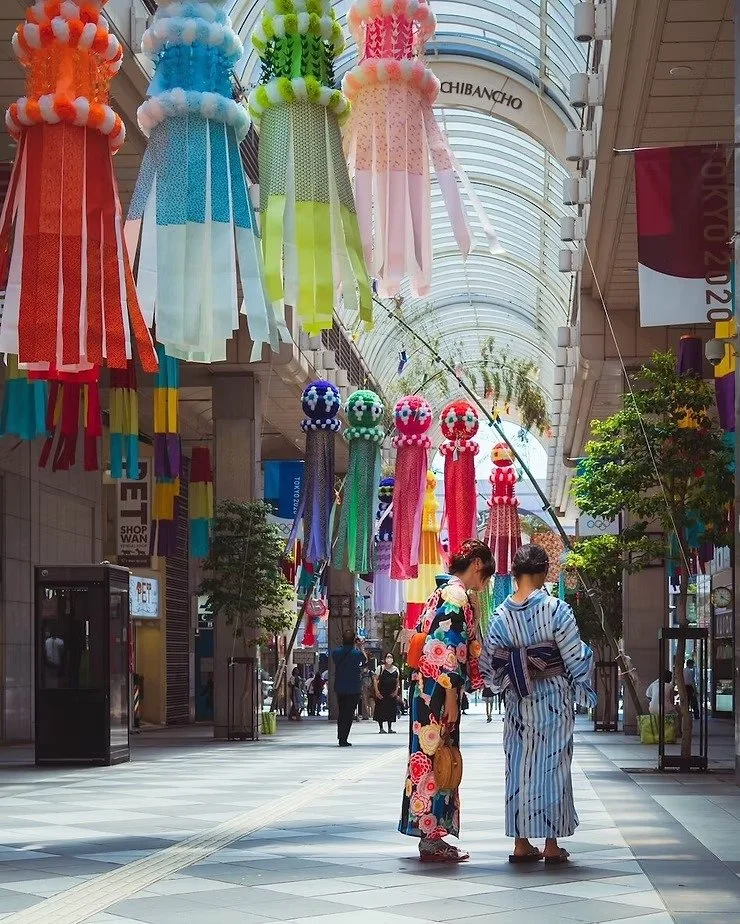 Two women in vibrant yukatas admire something on the street under colorful Tanabata streamers in a covered shopping arcade in Tokyo.