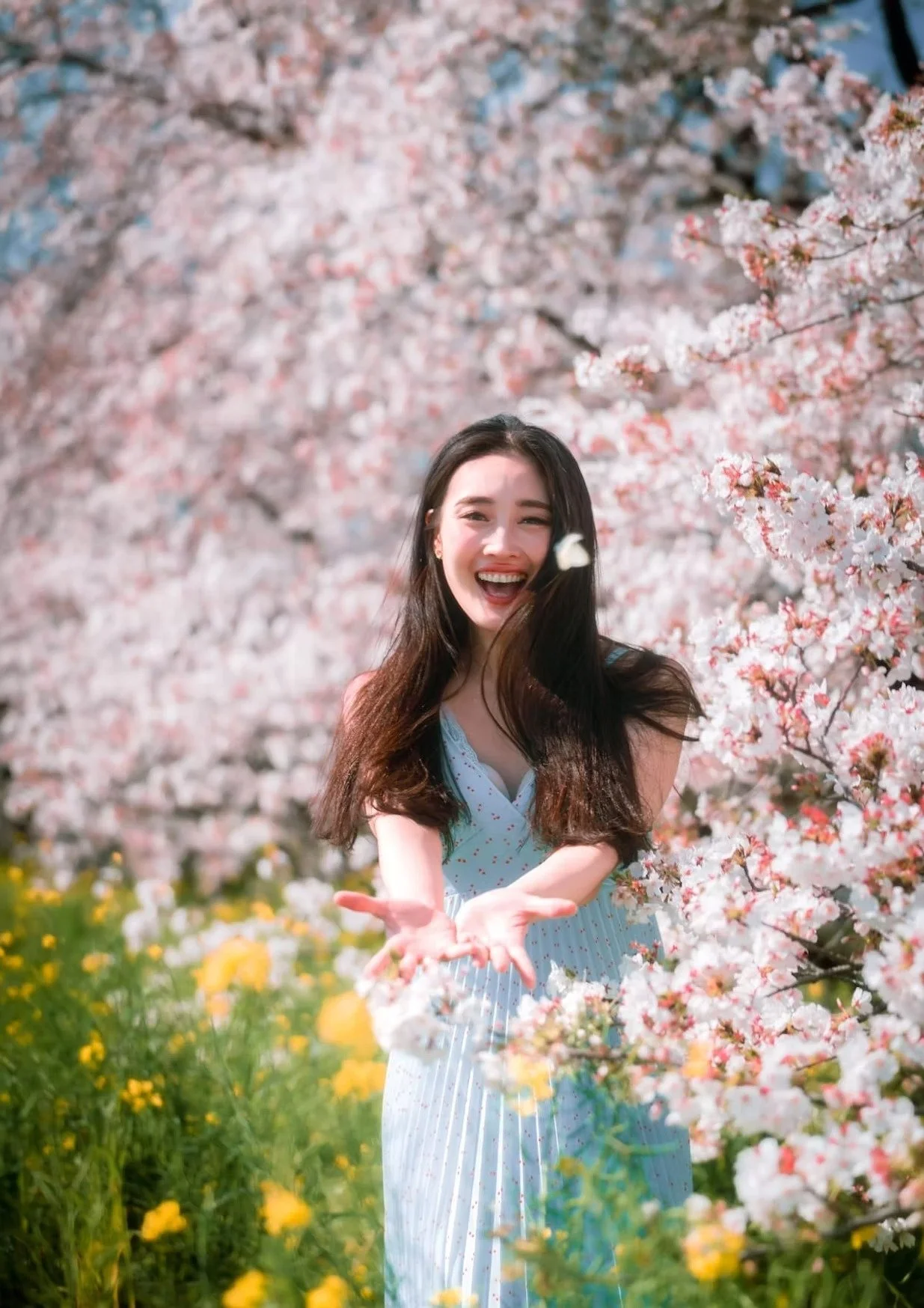A woman in a light blue dress smiles joyfully as she stands among cherry blossom trees in full bloom. Yellow flowers grow at the base, adding to the vibrant, springtime atmosphere.