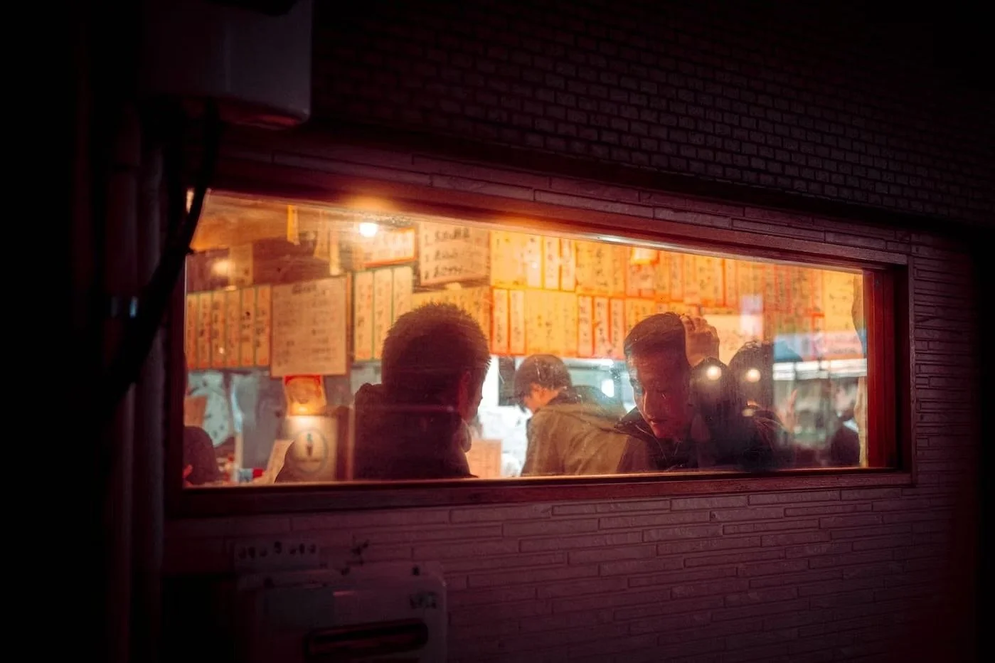 View through a restaurant window showing people inside, Japanese menu signs on the wall, warm lighting, and a brick exterior wall.