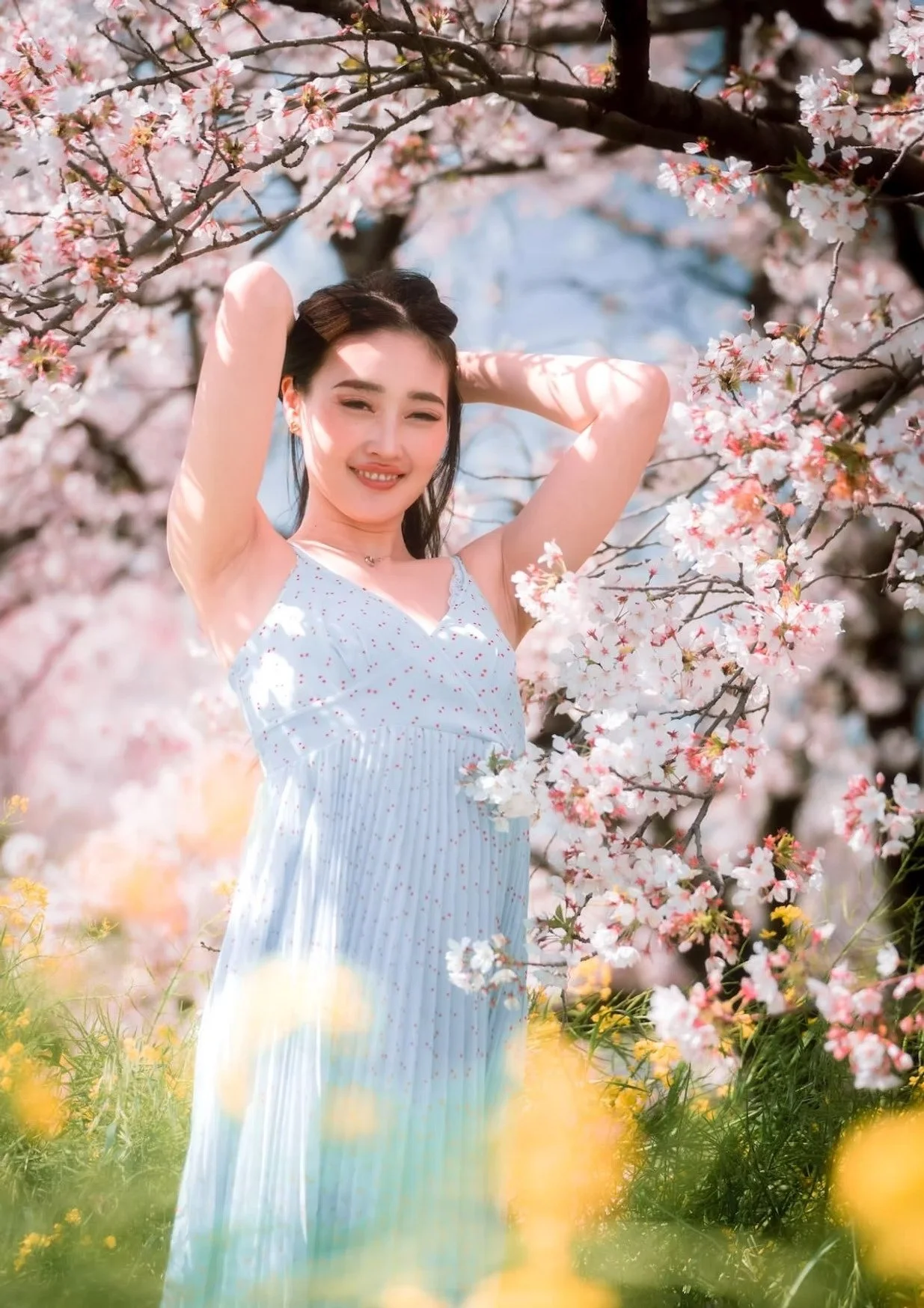 A woman in a light blue dress dances joyfully under the cherry blossom trees. Her arms are lifted, and she appears carefree, surrounded by pink flowers and yellow blooms in the grass, capturing the essence of a vibrant spring day.