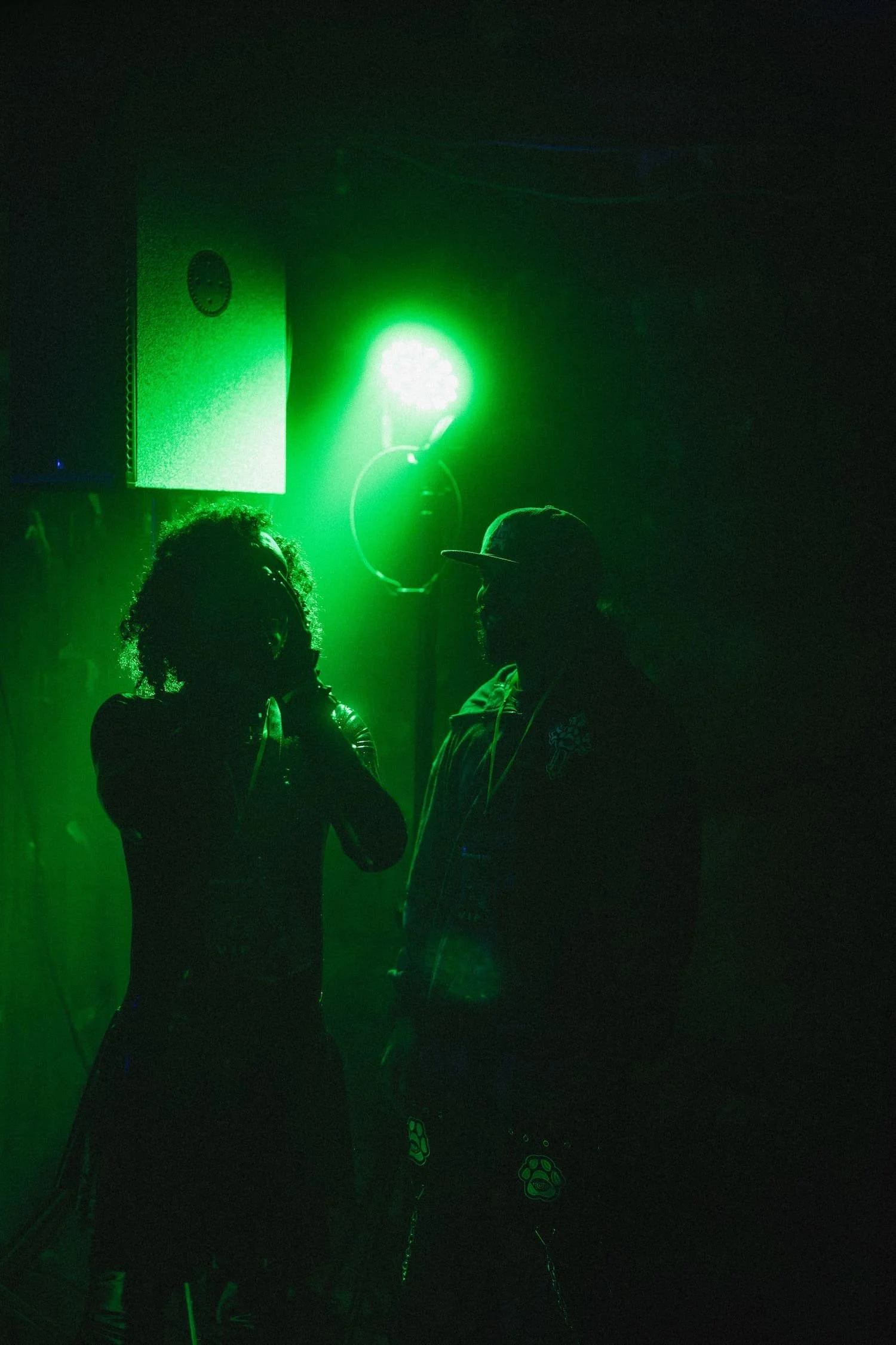 Bondage Ball 
---
Two people silhouetted against a dark background with green stage lighting, one person wearing glasses and the other wearing a cap.
