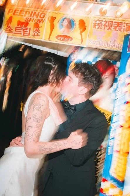 A couple embraces in the streets as they dance under the glowing lights, surrounded by neon signs. The photo captures a spontaneous and joyful moment with motion blur, highlighting the couple’s connection amidst the vibrant city scene.
