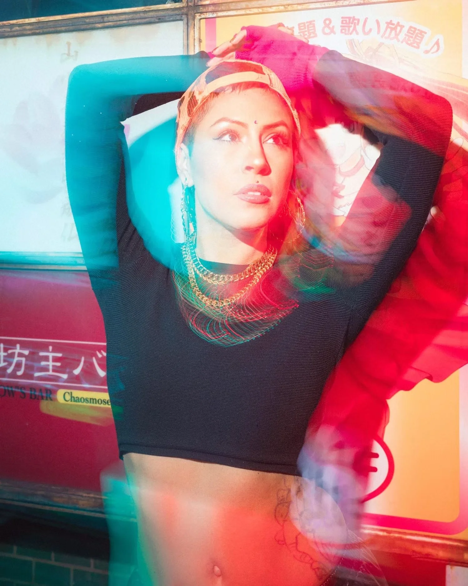 A woman with her hands behind her head, wearing a black top, earrings, a necklace, and a headscarf, posing in front of a colorful background with Japanese characters and some English text.
