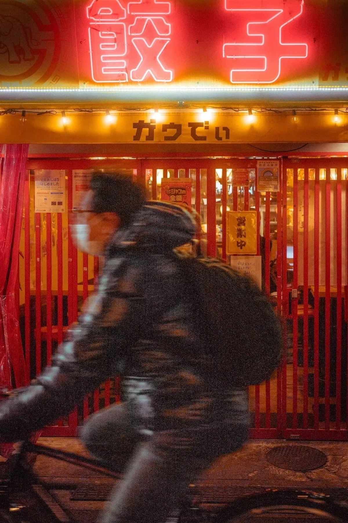 A man riding a bicycle past a brightly lit storefront with red vertical bars and neon signs in Japanese characters.