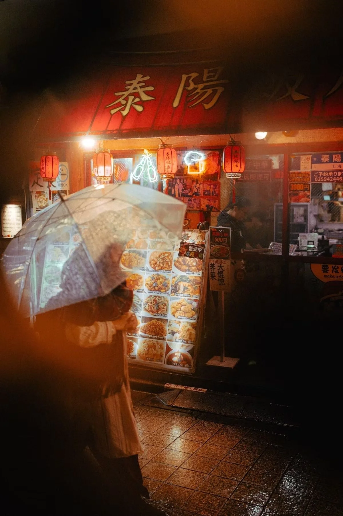 A person holding an umbrella while looking at a brightly lit menu board outside a restaurant at night, with neon signs and red lanterns hanging above in Nakano area.