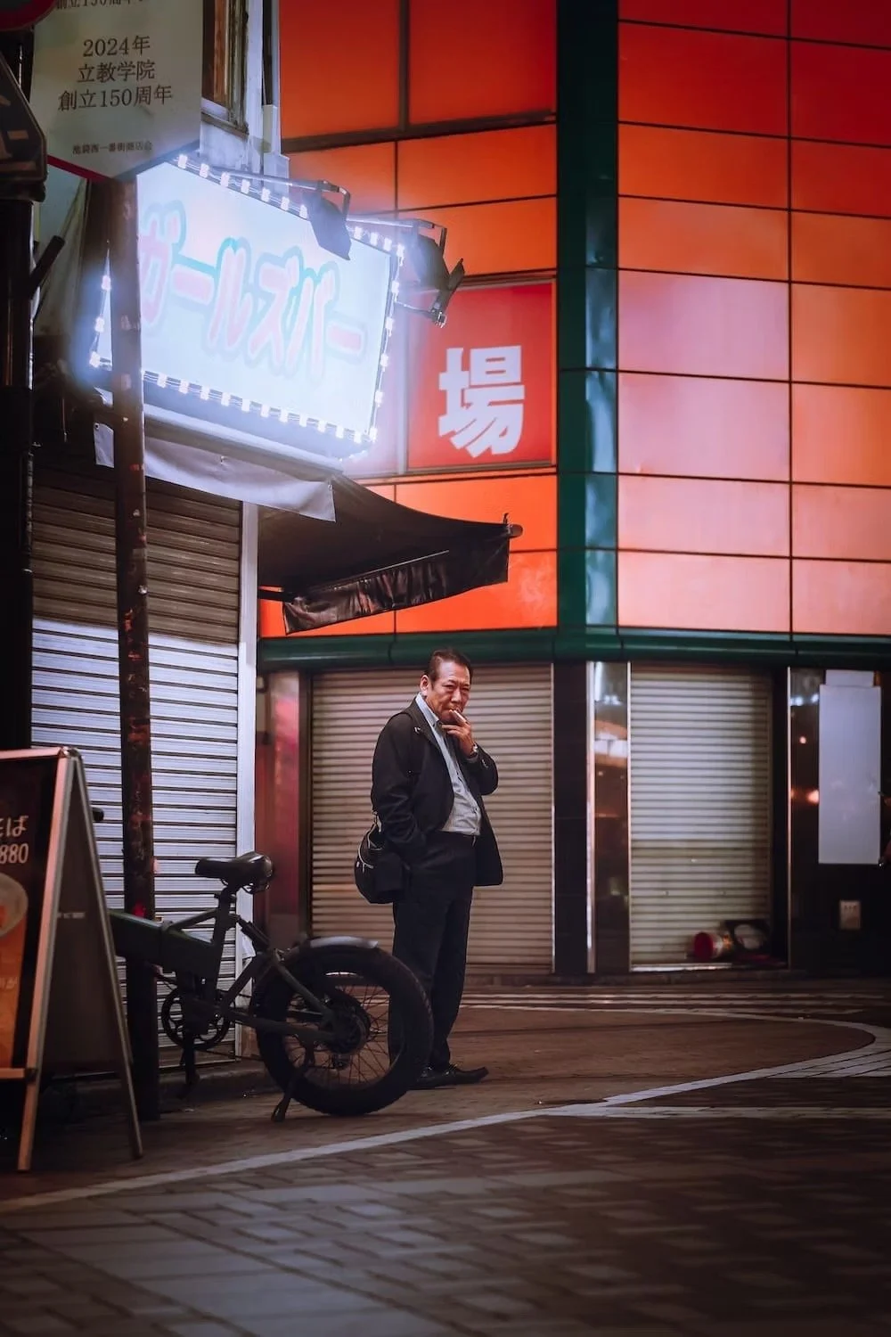 A Japanese salaryman in a suit standing on a city street at night next to a bicycle, with a large illuminated sign and a building with closed shutters behind him.