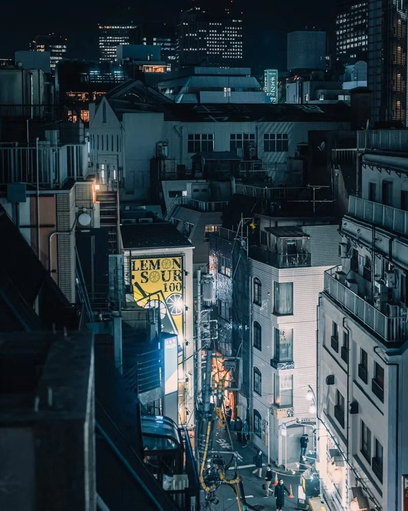 Night view of a city street with tall buildings, neon signs, and people walking on the sidewalk.