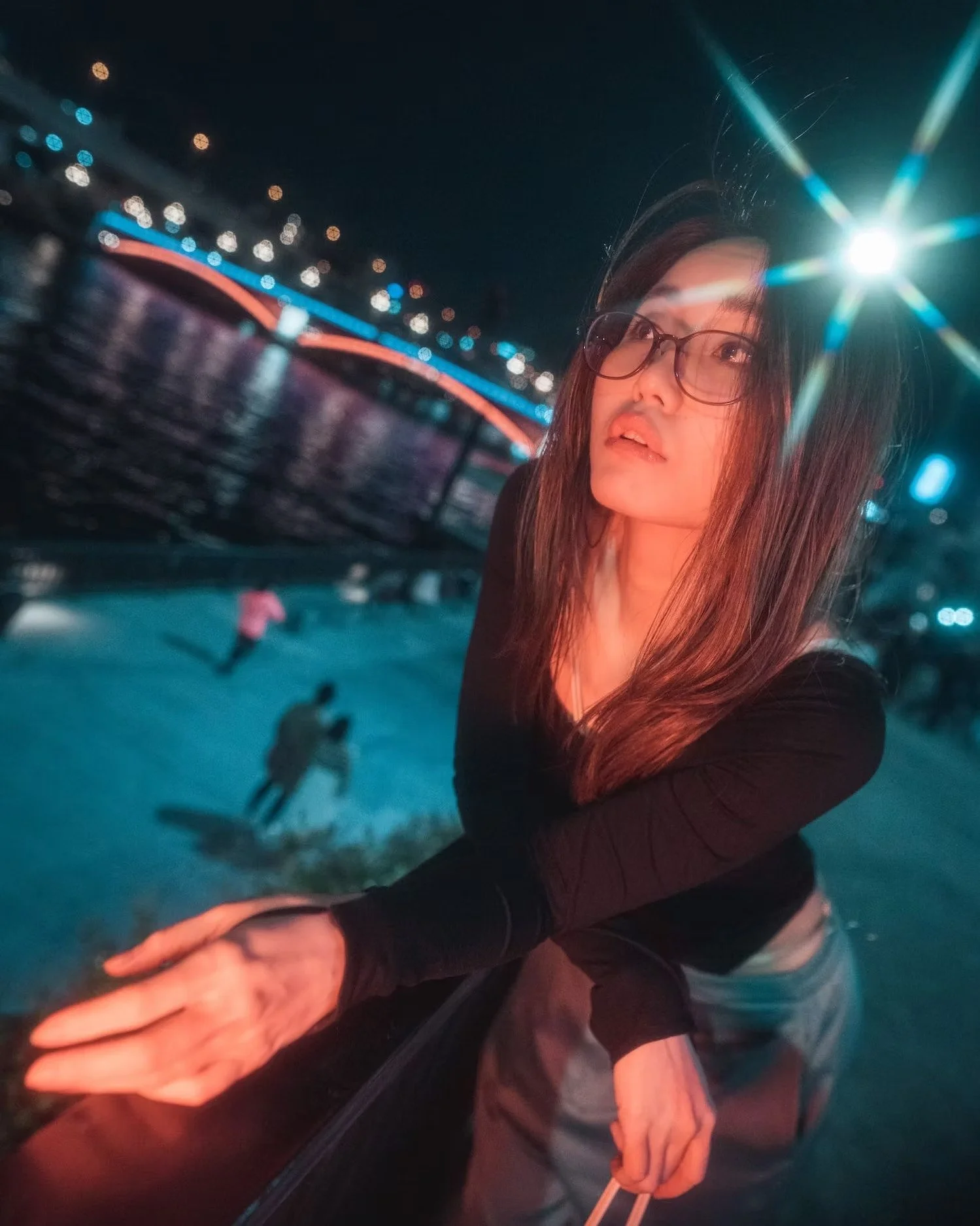 A woman with long hair and glasses standing outdoors at night, with ice skating rink and colorful buildings or bridge with lights in the background.