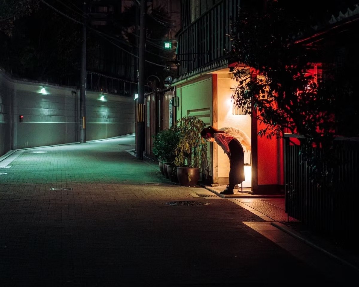 A person standing at the entrance of a building illuminated by warm light at night, with plants nearby, on a quiet city street in Kyoto, Japan shot by Tokyo street photographer Deniz Demir.