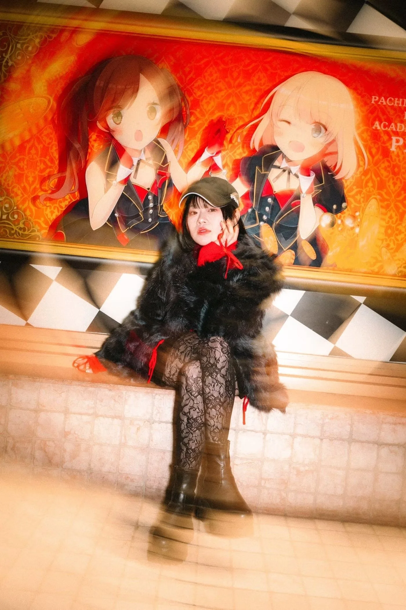 A Japanese woman dressed in a black fur coat, red scarf, and patterned black tights, sitting on a ledge with her legs crossed, in front of a colorful anime-themed poster featuring two animated girls in black and red outfits. Shot by Tokyo portrait ph