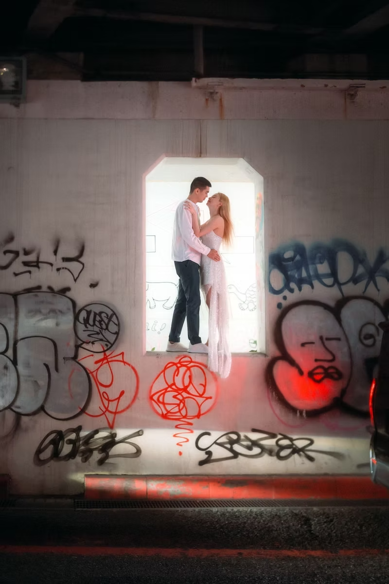 A couple stands in front of a graffiti-covered wall at night. The man lifts the woman in a romantic pose as they are illuminated by soft light, adding a cool urban vibe to the scene.