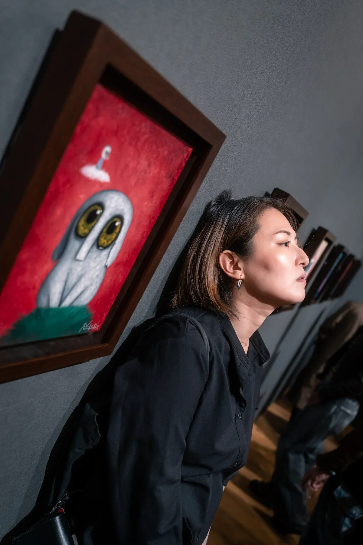 August Vilella
---
Creative editorial portrait of a woman in black posing beside a framed surrealist painting featuring a white creature with oversized blue eyes at August Vilella's contemporary art exhibition.
