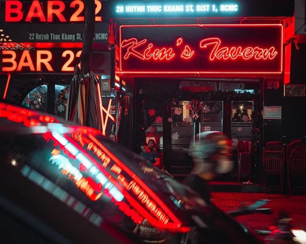 Night view of Kim's Tavern with bright red neon signs, people sitting inside, and a blurred motorcycle passing by outside. Shot by Tokyo street photographer Deniz Demir on the streets of Saigon Vietnam.