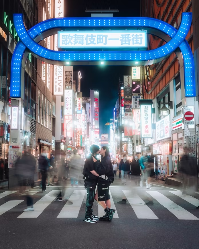 couple kisses on the crosswalk beneath the "I Love Kabukicho" neon archway. The busy Kabukicho street in Tokyo is filled with colorful neon signs and a blurred crowd of pedestrians, capturing the vibrant energy of the city at night