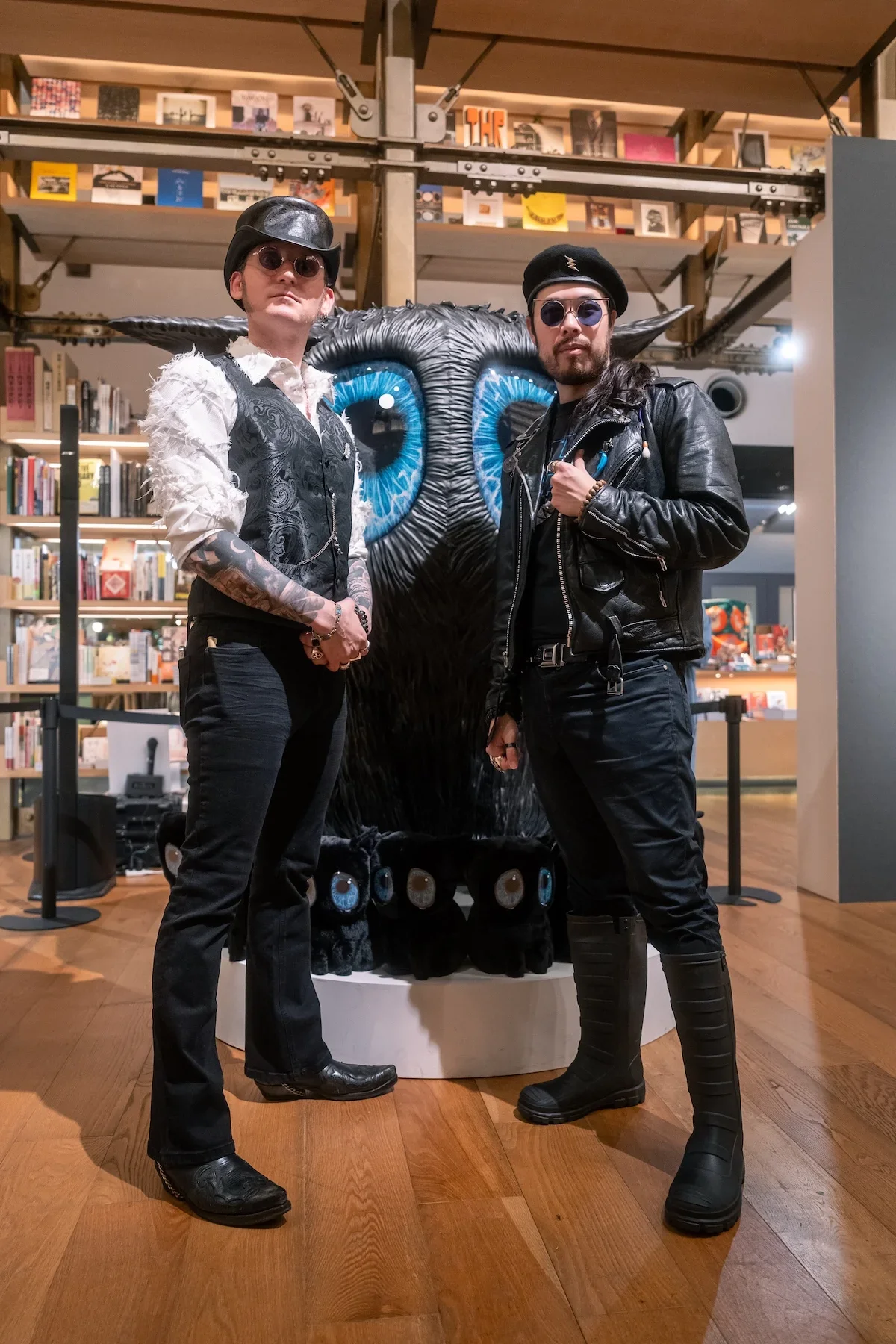August Vilella
---
Surrealist artist August Vilella, wearing his signature hat, round glasses, and tattooed arms, poses alongside a guest dressed in black leather in front of a large blue-eyed creature sculpture at his contemporary art exhibition in 