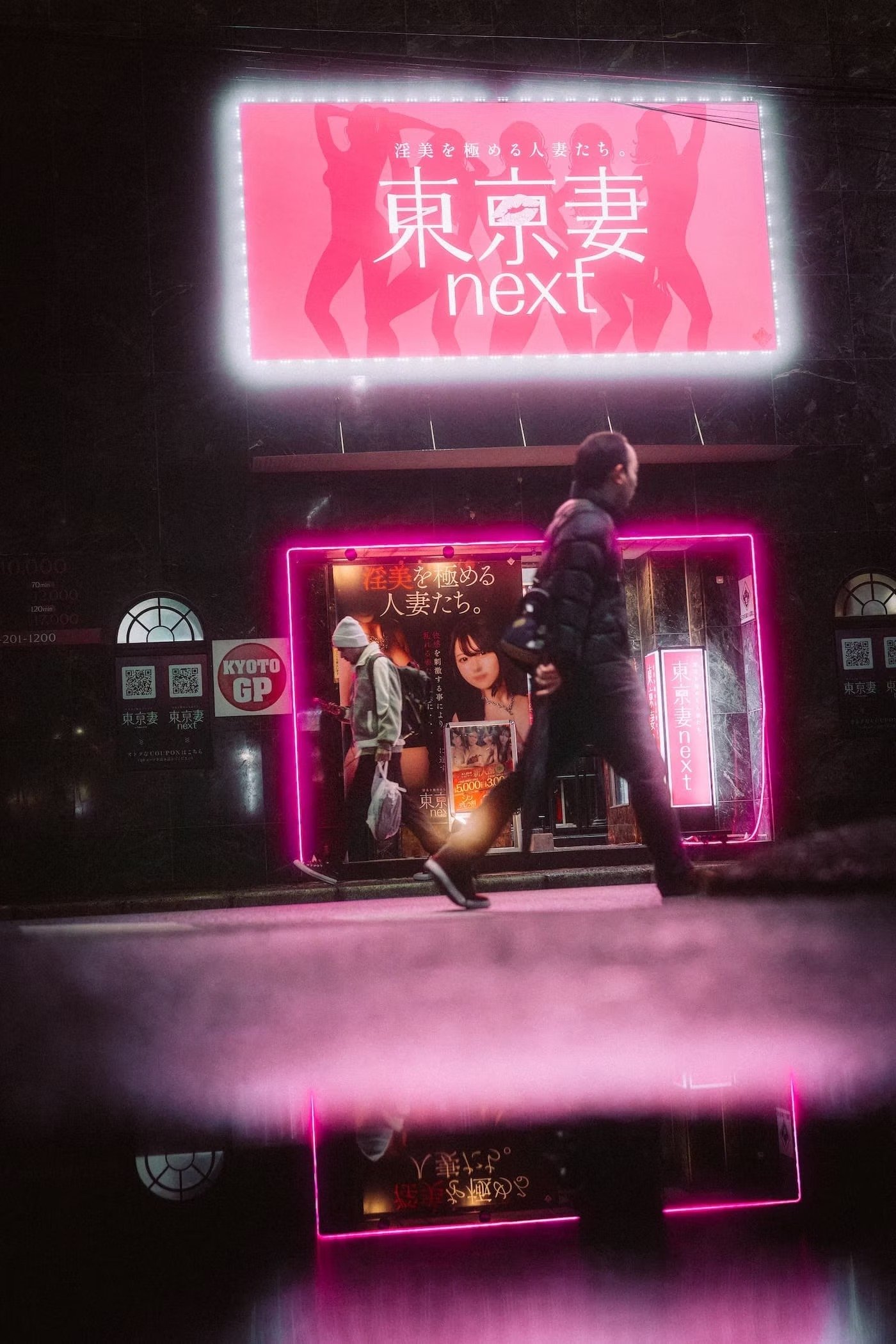 Neon pink sign in Japanese with the word 'next' in English, featuring silhouettes of women on a dark wall and a person walking by in the foreground. Shot by Tokyo street photographer Deniz Demir.