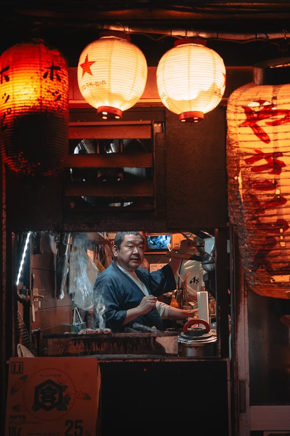 A man working at a yakitori food stall inside a Japanese restaurant or street food vendor, illuminated by traditional paper lanterns with Japanese characters and symbols hanging above. Shot by Tokyo street photographer Deniz Demir.
