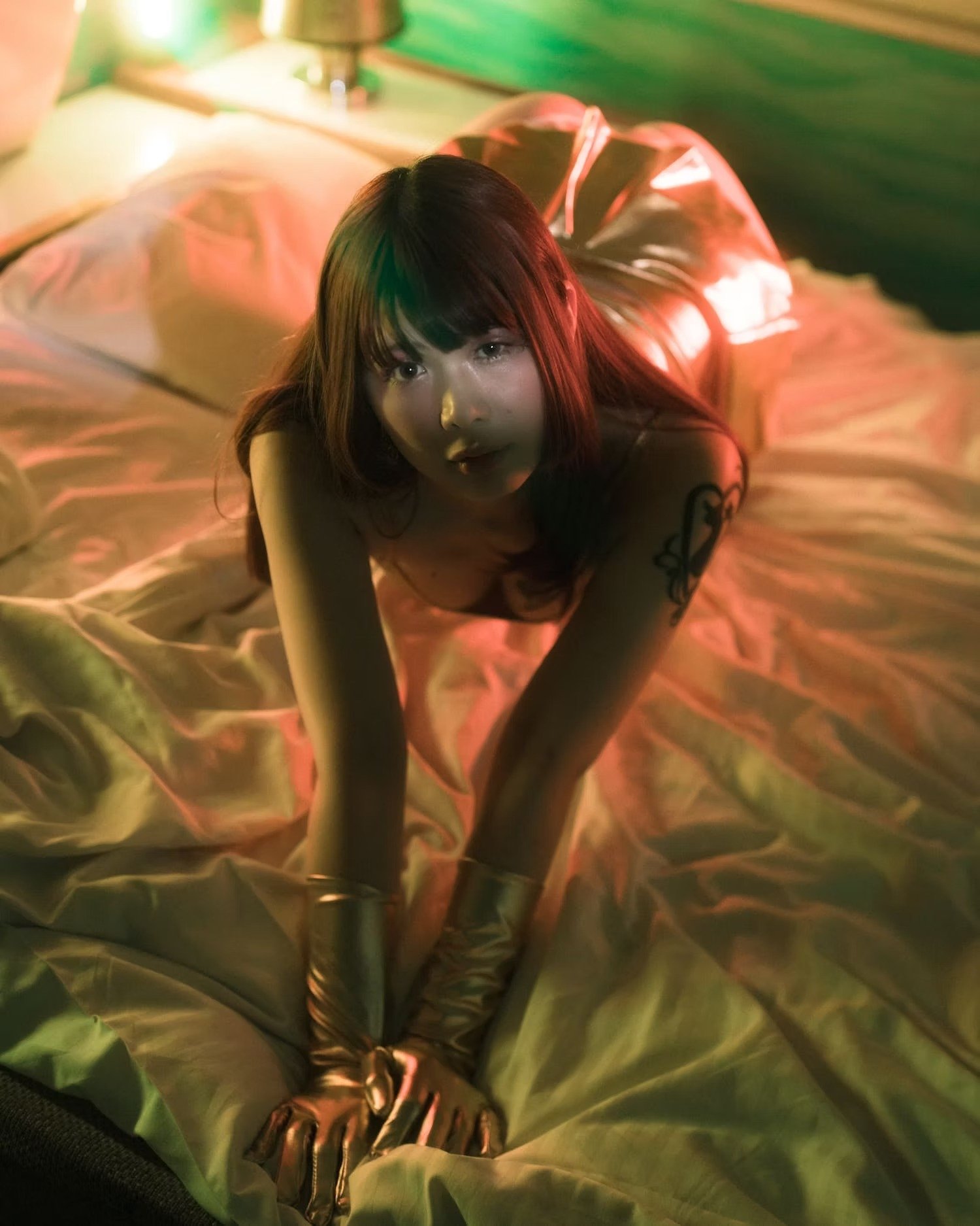 A woman with long dark hair and tattoos on her arm, lying on a bed with shiny, metallic-looking sheets, illuminated by colorful, ambient lighting.