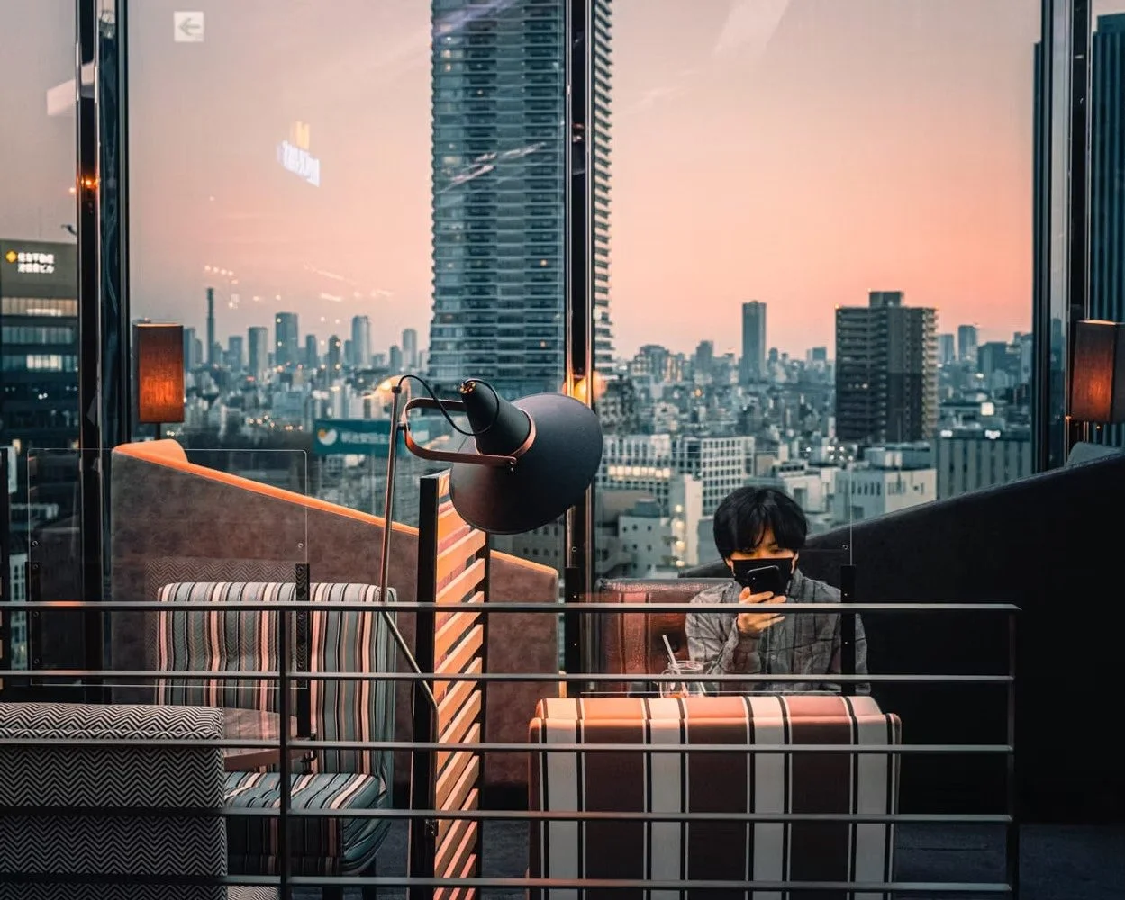A person wearing a face mask sitting at a table in a modern indoor space with large floor-to-ceiling glass windows showing a city skyline at sunset. The person is looking at a smartphone.