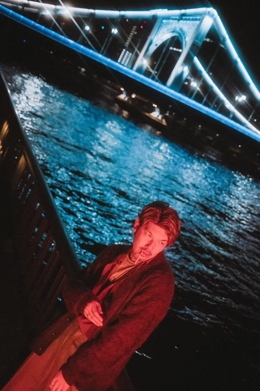 A young man with styled hair and a cigarette in his mouth standing on a waterfront at night, with a brightly lit suspension bridge in the background reflecting on the water. In Tokyo shot by Tokyo portrait photographer Deniz Demir.