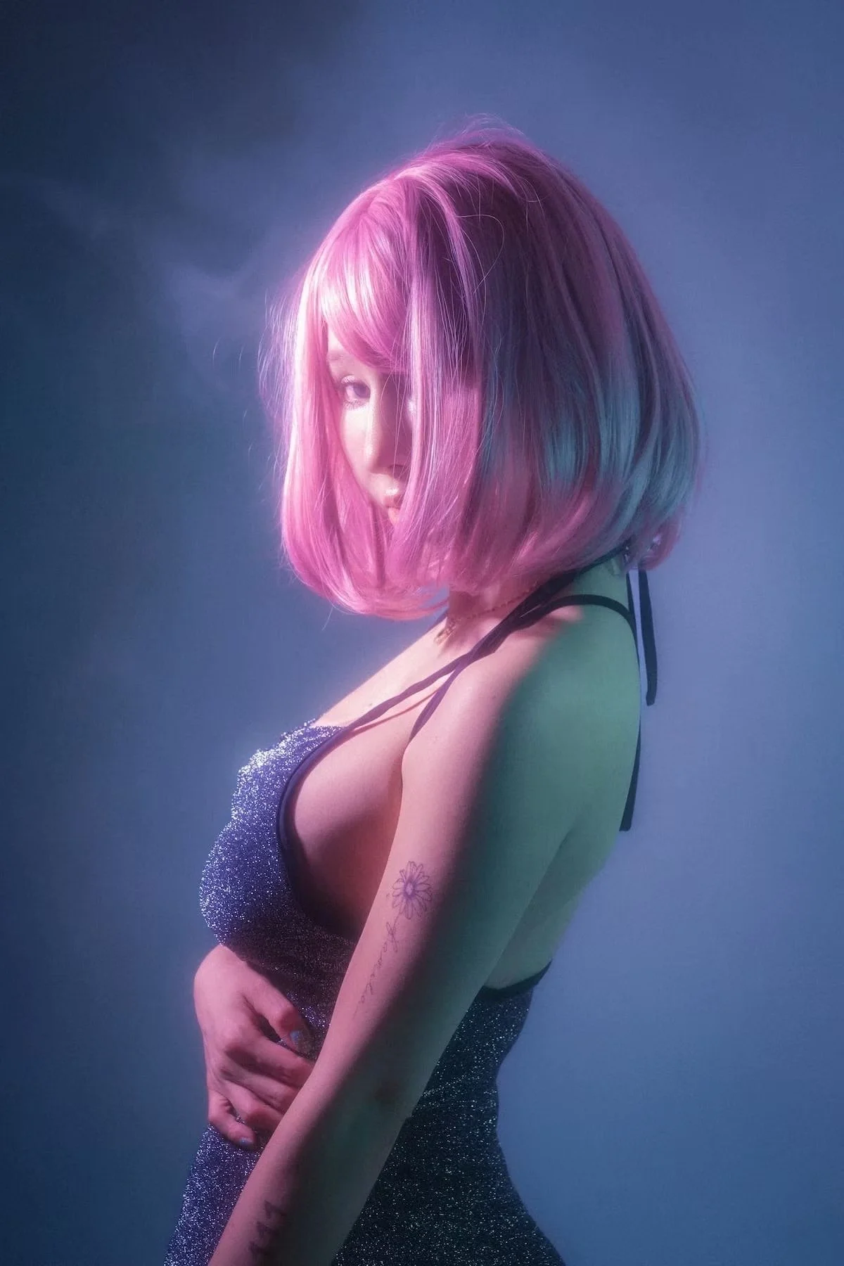 A woman with shoulder-length pink hair, wearing a sparkly black top and black straps on her shoulders, is posing with her body turned to the side and her face partially obscured by her hair against a blue background.