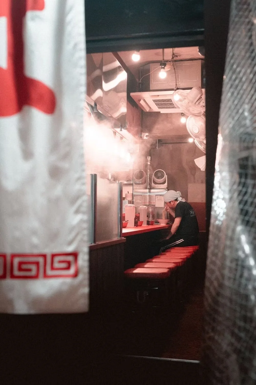 A person sitting alone at a small counter in a dimly lit Japanese Ramen restaurant, wearing a white head covering and black clothing, surrounded by hanging light bulbs and kitchen equipment.