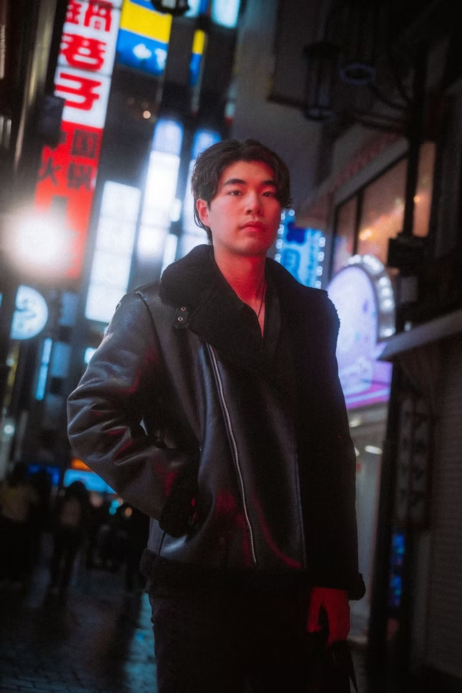 A young man wearing a black leather jacket and orange sneakers stands in a brightly lit street of Tokyo, illuminated by neon signs.
