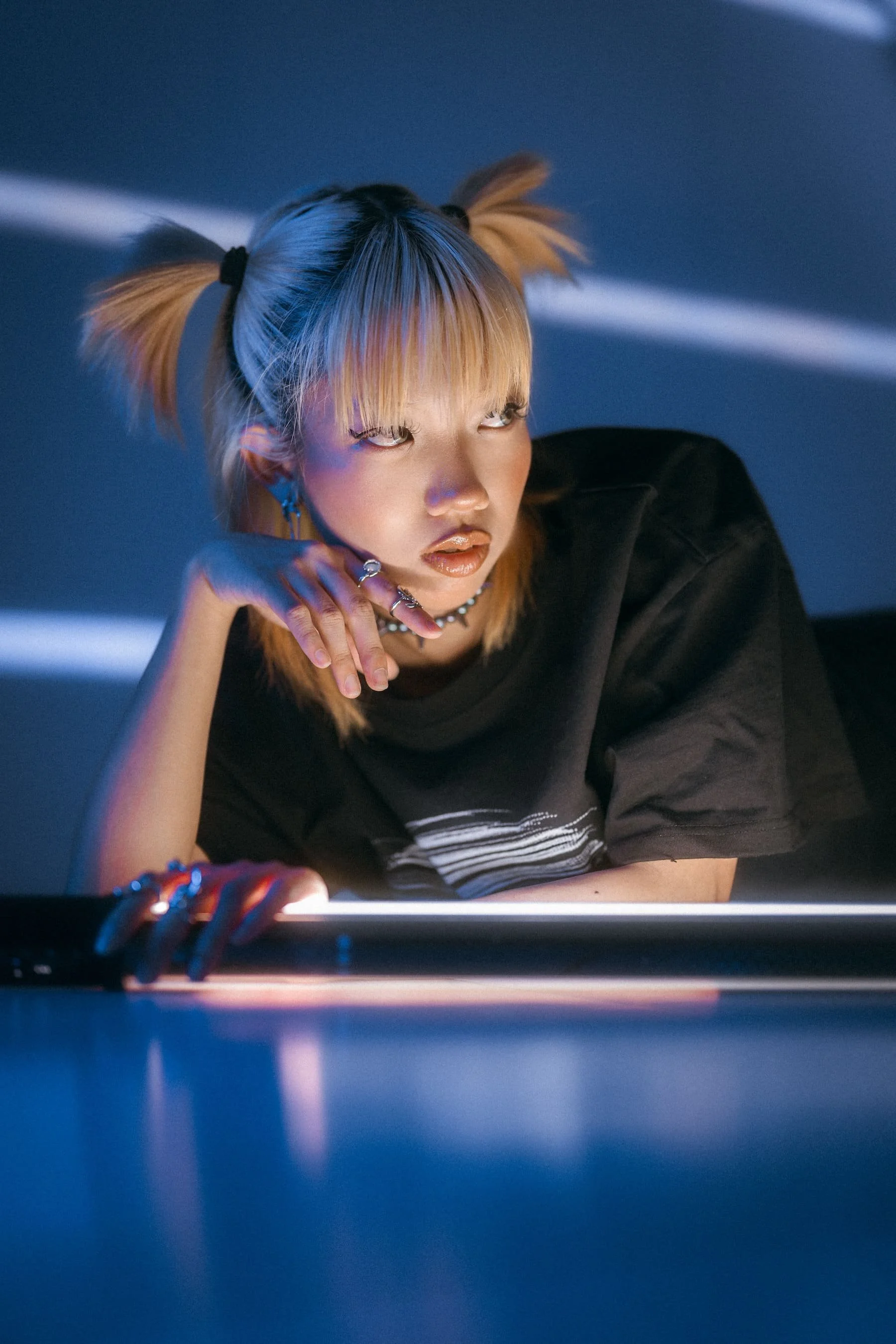 A young woman with blonde hair in pigtails and bangs, wearing a black T-shirt with striped detail, jewelry, and makeup, looking at a screen or reflective surface with a focused expression in a dimly lit environment with blue tones.