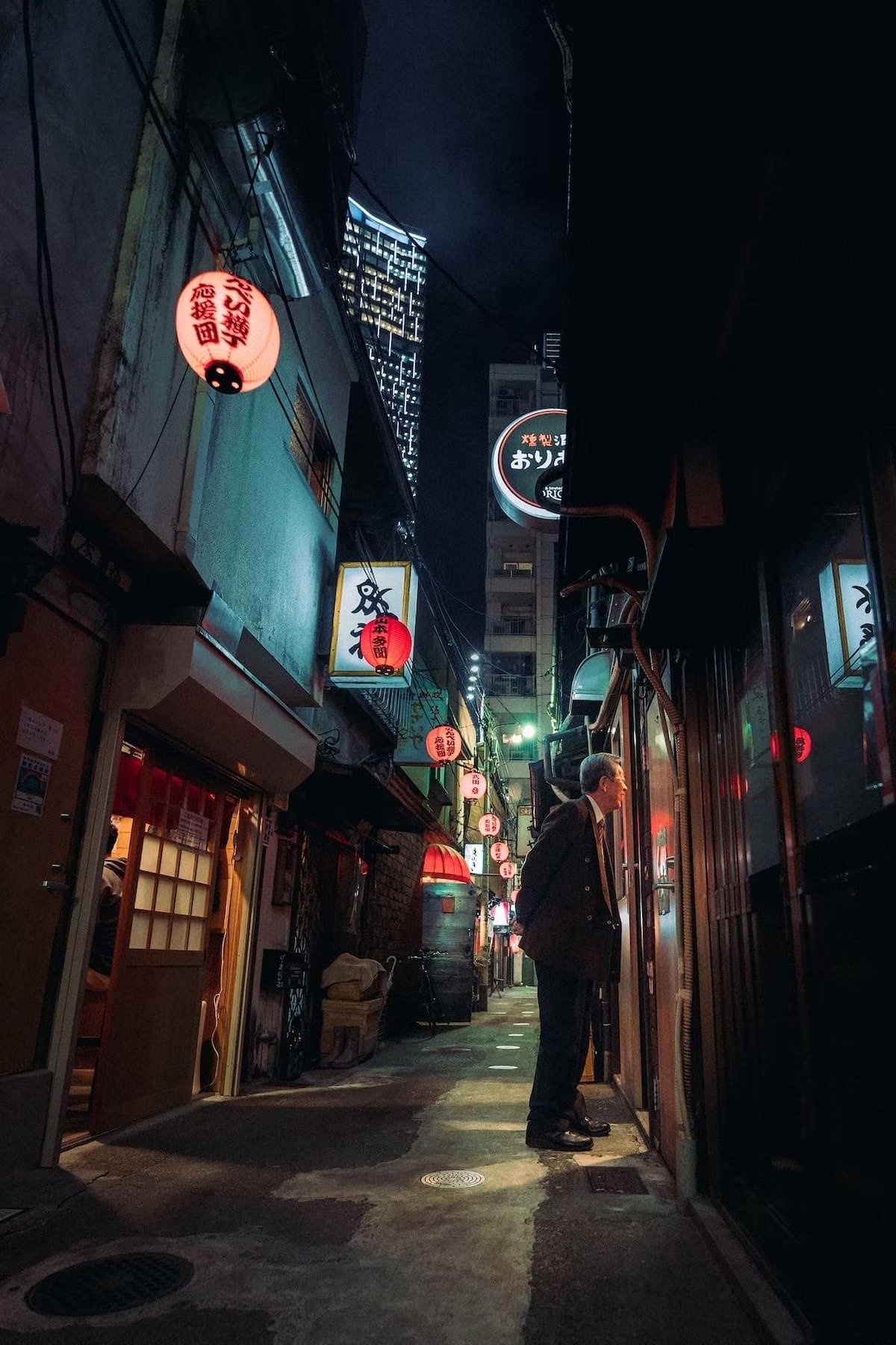 A narrow, illuminated alleyway in an urban area at night, with hanging paper lanterns with Japanese characters, small storefronts on either side, and a salaryman in a suit standing outside one of the shops in Shibuya.