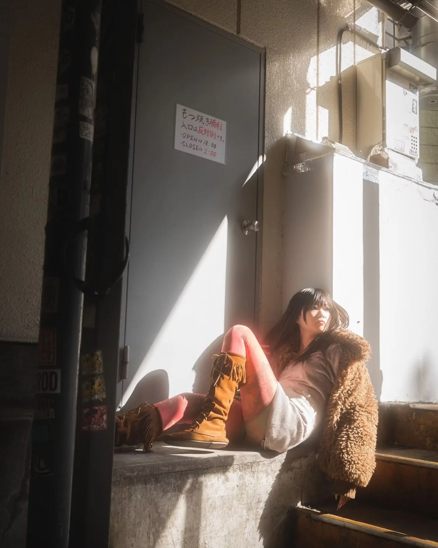 A woman sitting on a concrete ledge with sunlight shining on her, wearing a brown furry coat and rust-colored boots, with another person's legs stretched out in front of her and a sign on the wall nearby. The sign appears to have text in Japanese and