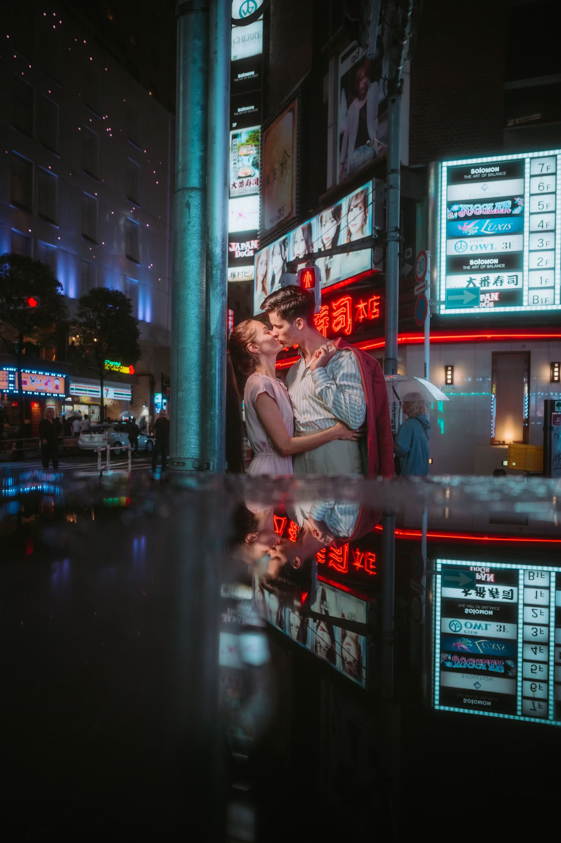 A young couple stands close and shares a kiss beneath street lights and neon signs in an urban nighttime setting. The scene reflects on a wet surface, creating a mirror image of their figures and the city lights.