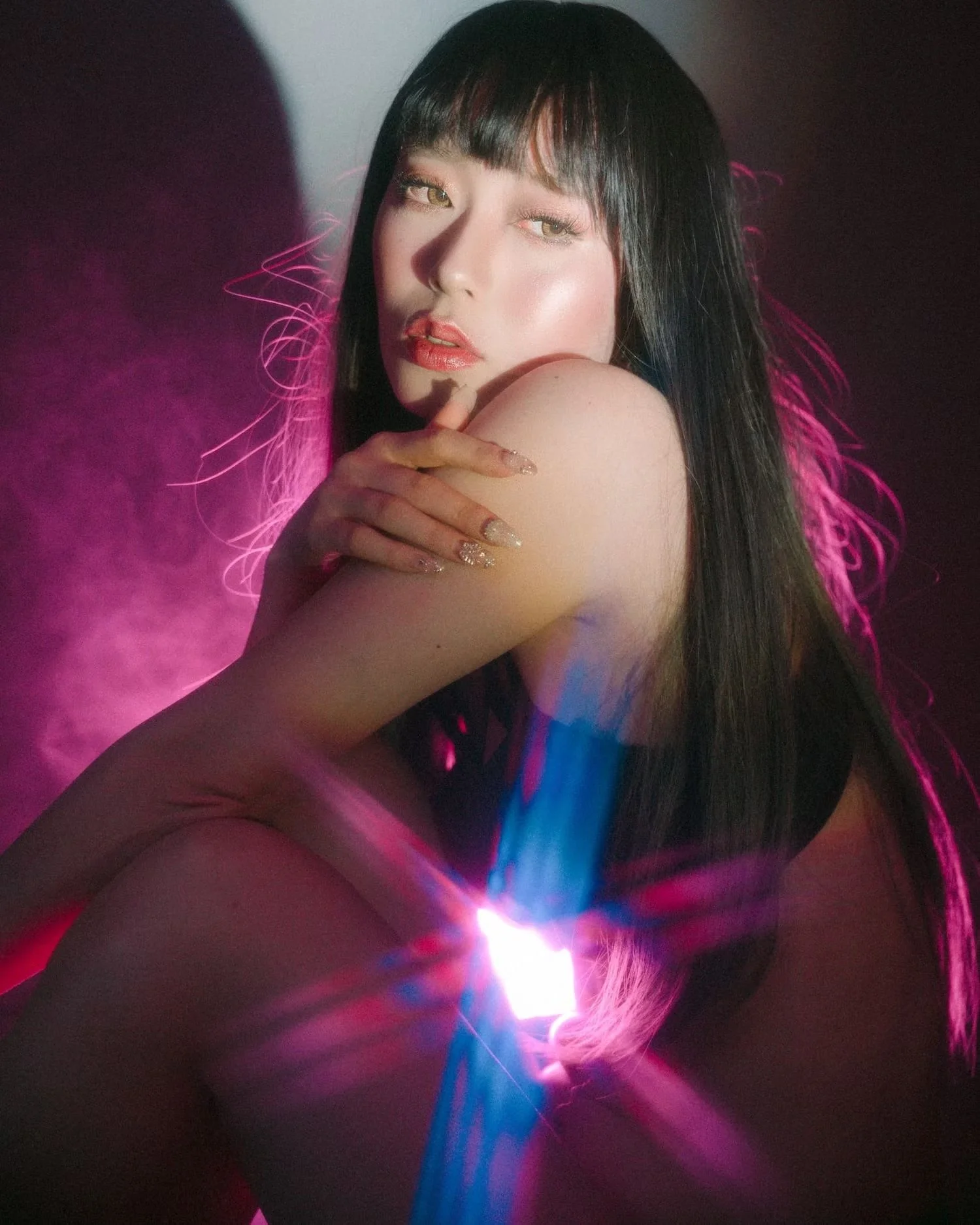 A woman with long black hair, featuring blunt bangs, posing with her hand on her shoulder. She has glossy lips, eye makeup, and is illuminated by colorful neon lights in pink, purple, and blue.