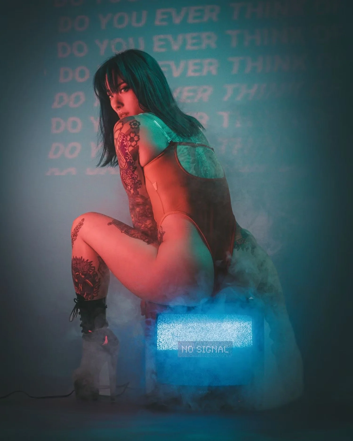 A woman with dark hair and tattoos on her arm and leg kneeling, wearing a tank top and shorts. Behind her, a projected screen displays the repeated text 'DO YOU EVER THIN' and a message at the bottom reads 'NO SIGNAL'.