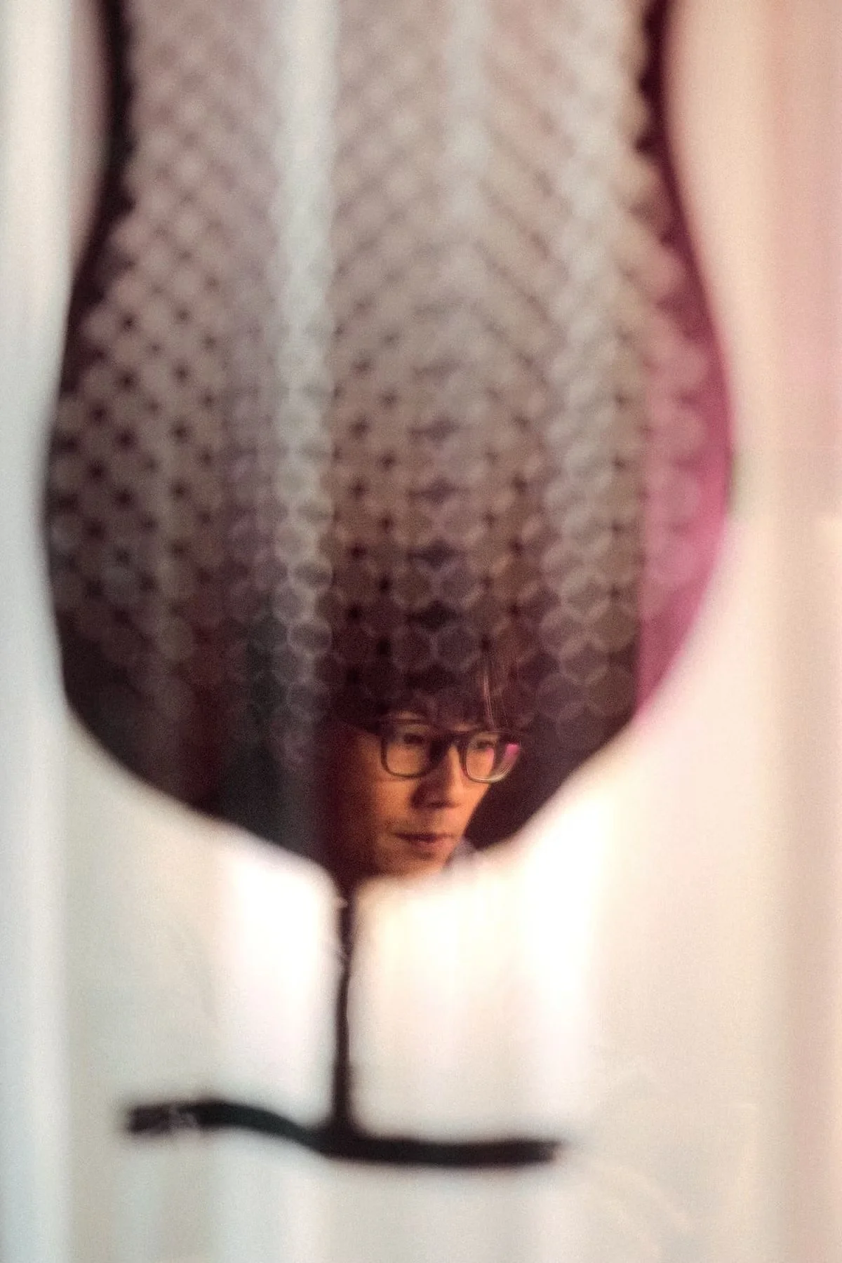 Reflection of a person with glasses and a hat, seen through an irregularly shaped mirror or window.