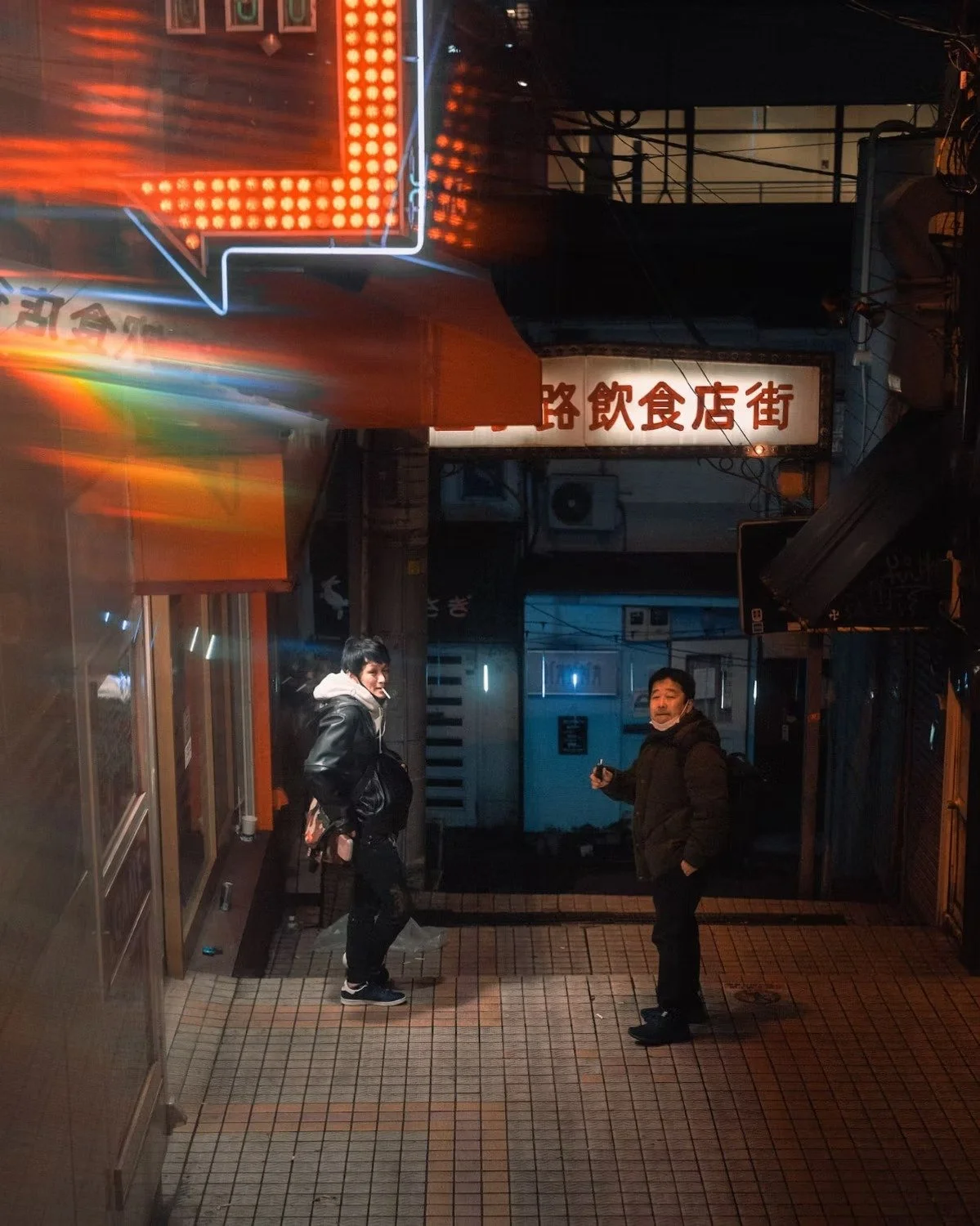 Two men standing in a narrow alley with neon signs in Japanese. One man is wearing a black jacket and smiling, the other is holding a phone and looking at the camera. Nighttime scene with urban street lighting.