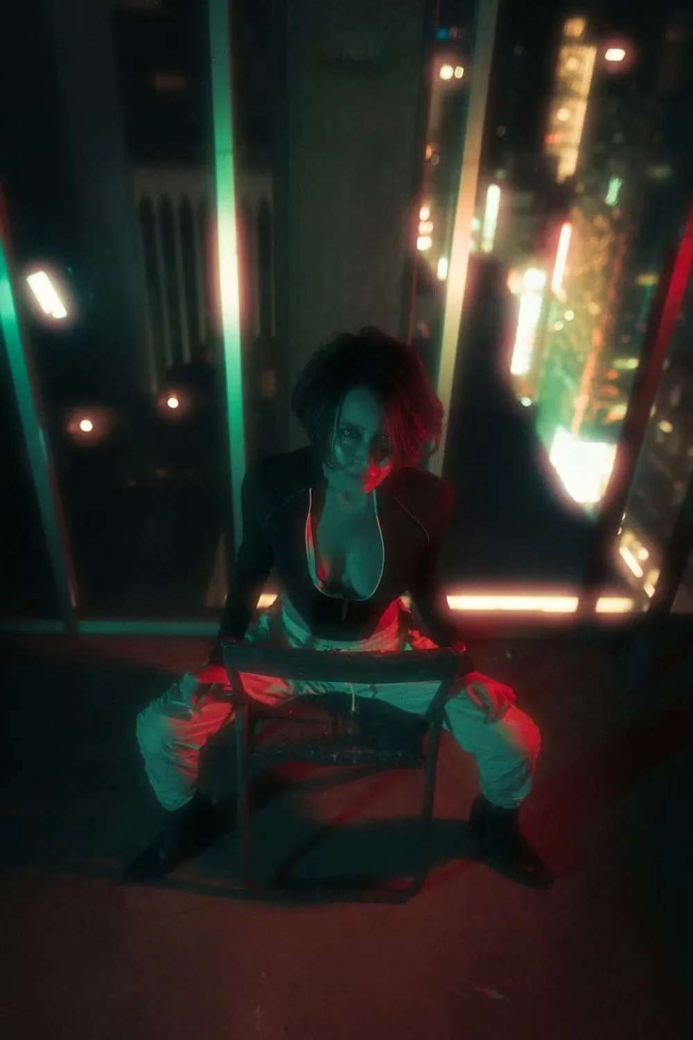 Person with short dark hair and in revealing clothing sitting on a chair in a dimly lit room with cityscape lights visible through windows behind them.
