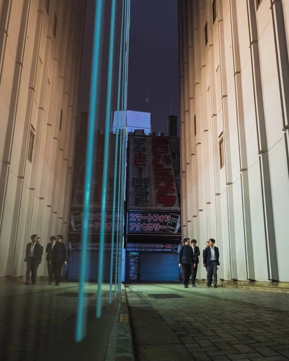 Nighttime street scene with a group of men in suits walking and talking near a tall building with advertising signs, illuminated with blue neon lights in a Tokyo alley evoking cyberpunk scenes.