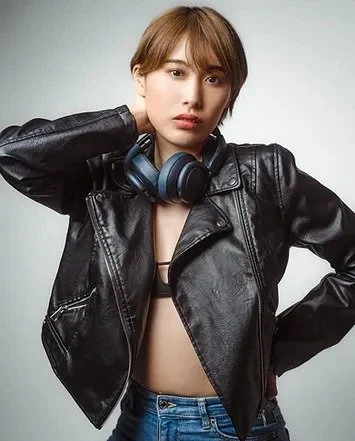 A young woman with short hair wearing a black leather jacket and headphones around her neck, posing with one hand behind her head against a plain background.