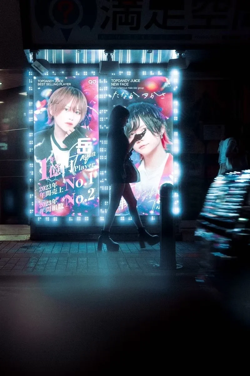 Two illuminated posters featuring a male idol, with a person walking in front of them, obscuring part of the images. The posters promote a top and juke idol group, highlighting achievements in 2023, with vibrant lights and colors shot in Kabukicho Sh