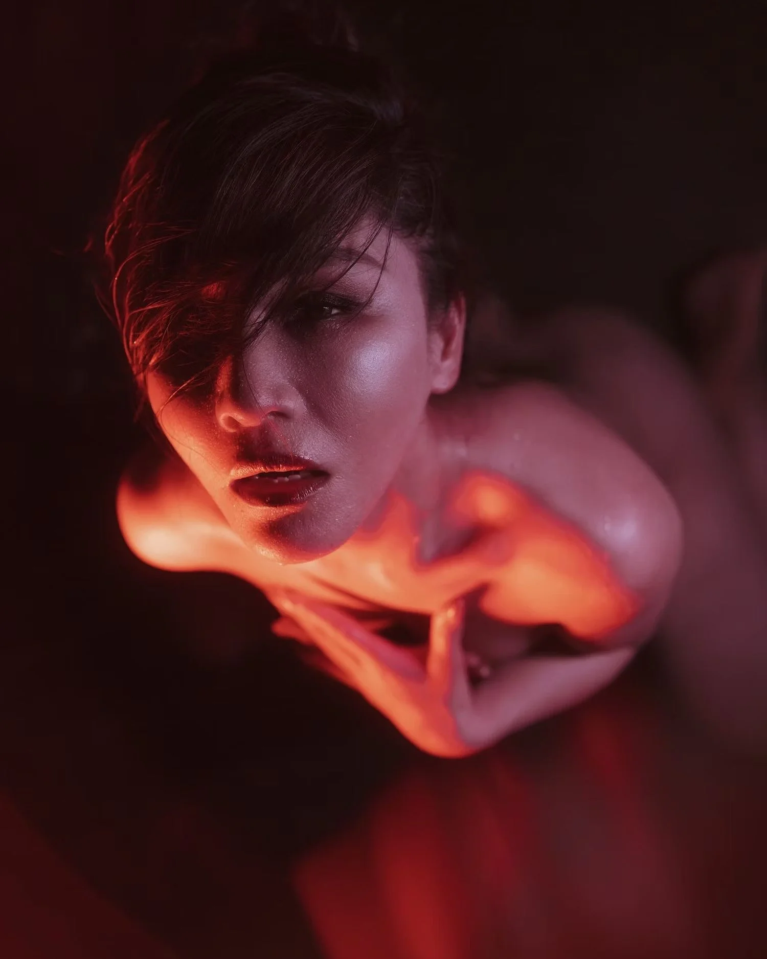 A person with short, dark hair, partially covering one eye, has their face illuminated by warm, red lighting, creating a dramatic, artistic effect.