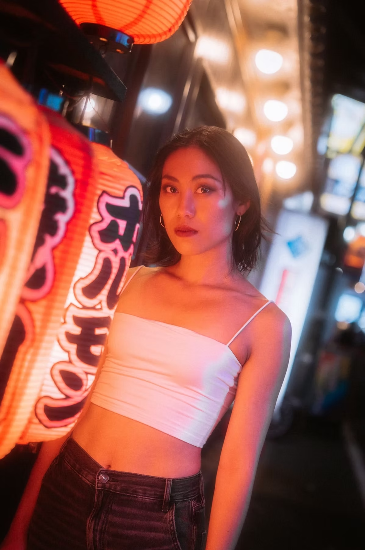 woman in a white crop top and black pants stands in front of a glowing sign in the bustling streets of Tokyo at night.