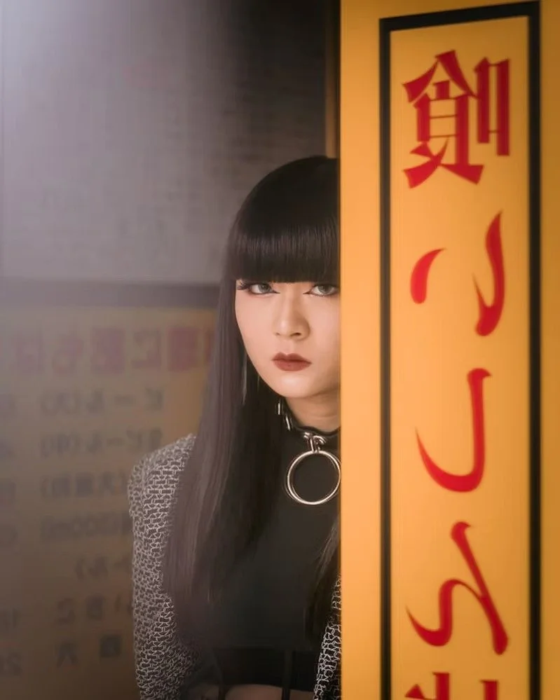 A woman with straight black hair and bangs peeks from behind a yellow and red sign, wearing a black choker with a large silver ring and a patterned jacket.